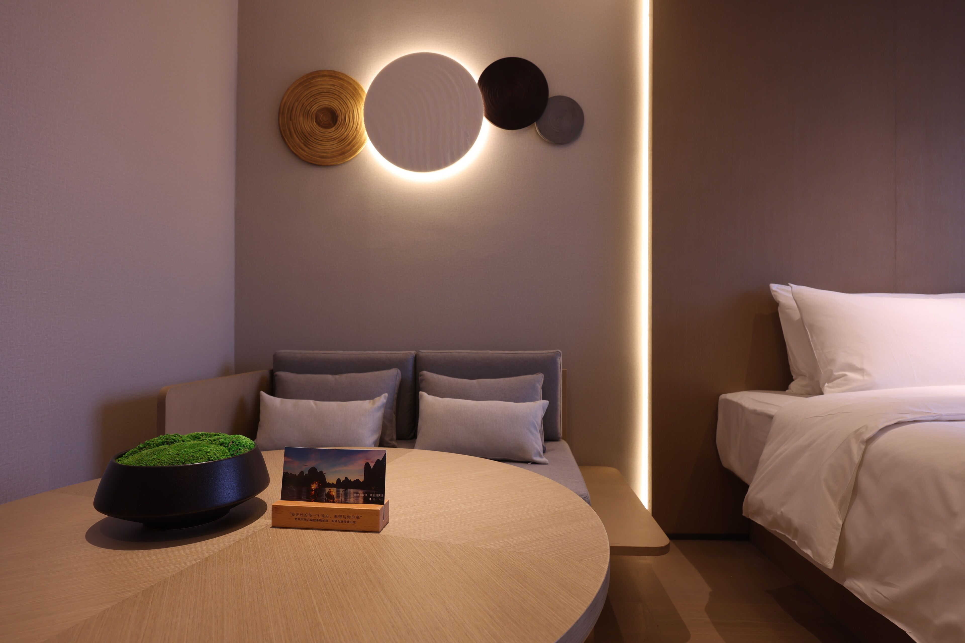 Een moderne hotelkamer met een ronde houten tafel, een kleine plant in pot, een lijst met een afbeelding en een bed met wit beddengoed.