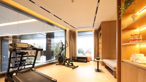 Un gimnasio con cinta de correr, elíptica y pesas, ventanas amplias y un estante de madera con toallas.