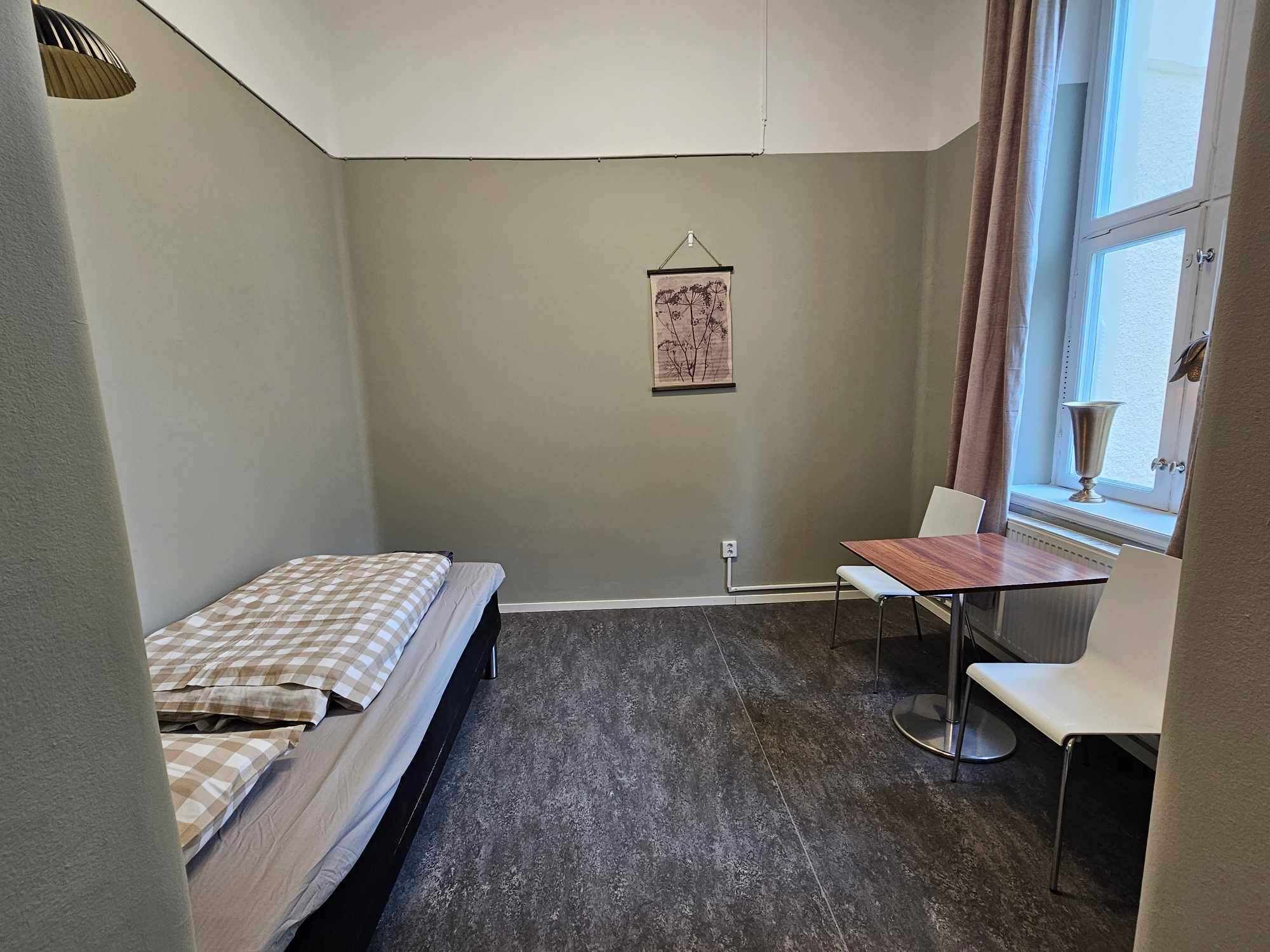 Een kleine, praktische kamer met een enkel bed, een tafel en een stoel. Er is een raam met gordijnen en een wandlamp.