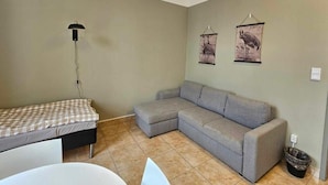 Una habitación con sofá gris, cama pequeña con manta a cuadros, lámpara de pared y dos cuadros decorativos.