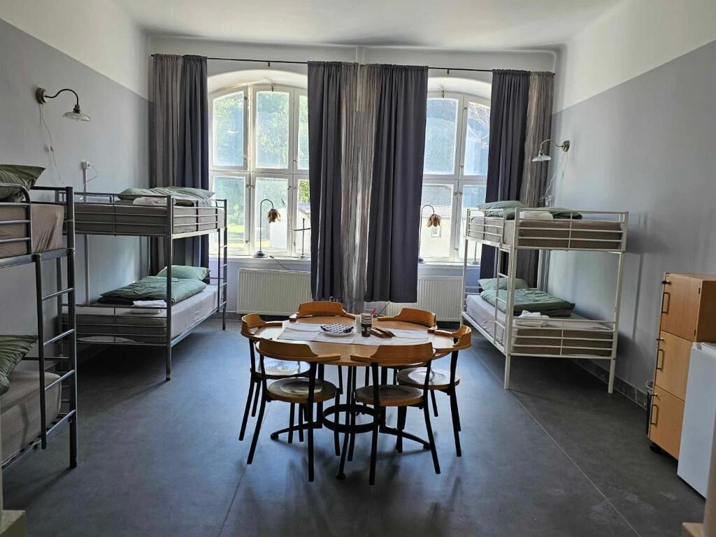 A dormitory room with bunk beds, a round table, chairs, and a radiator.