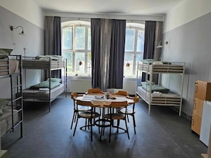 A dormitory room with bunk beds, a round table, chairs, and a radiator.