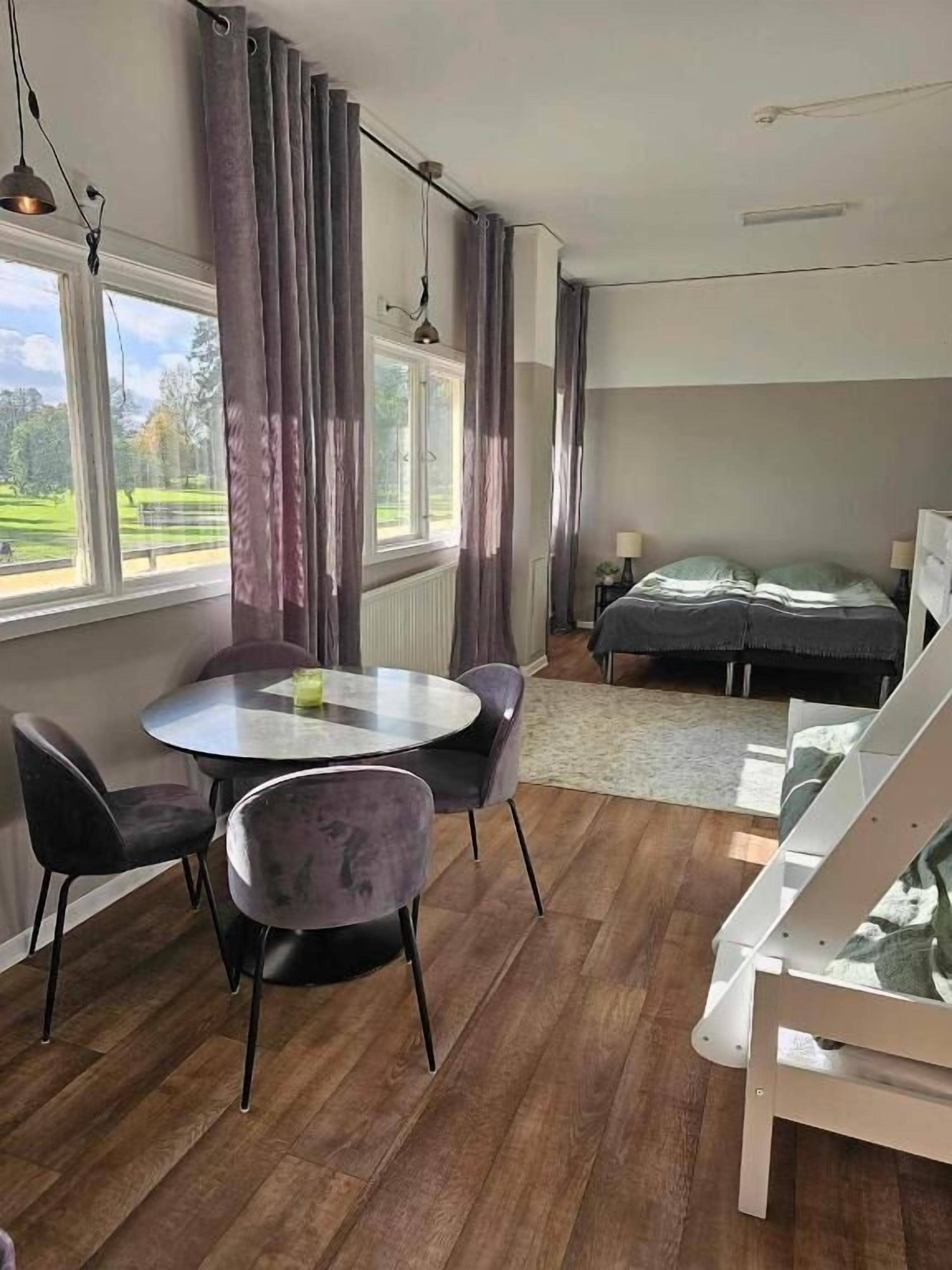 Una habitación con cama, una mesa redonda con sillas y vistas a un paisaje verde a través de amplios ventanales.