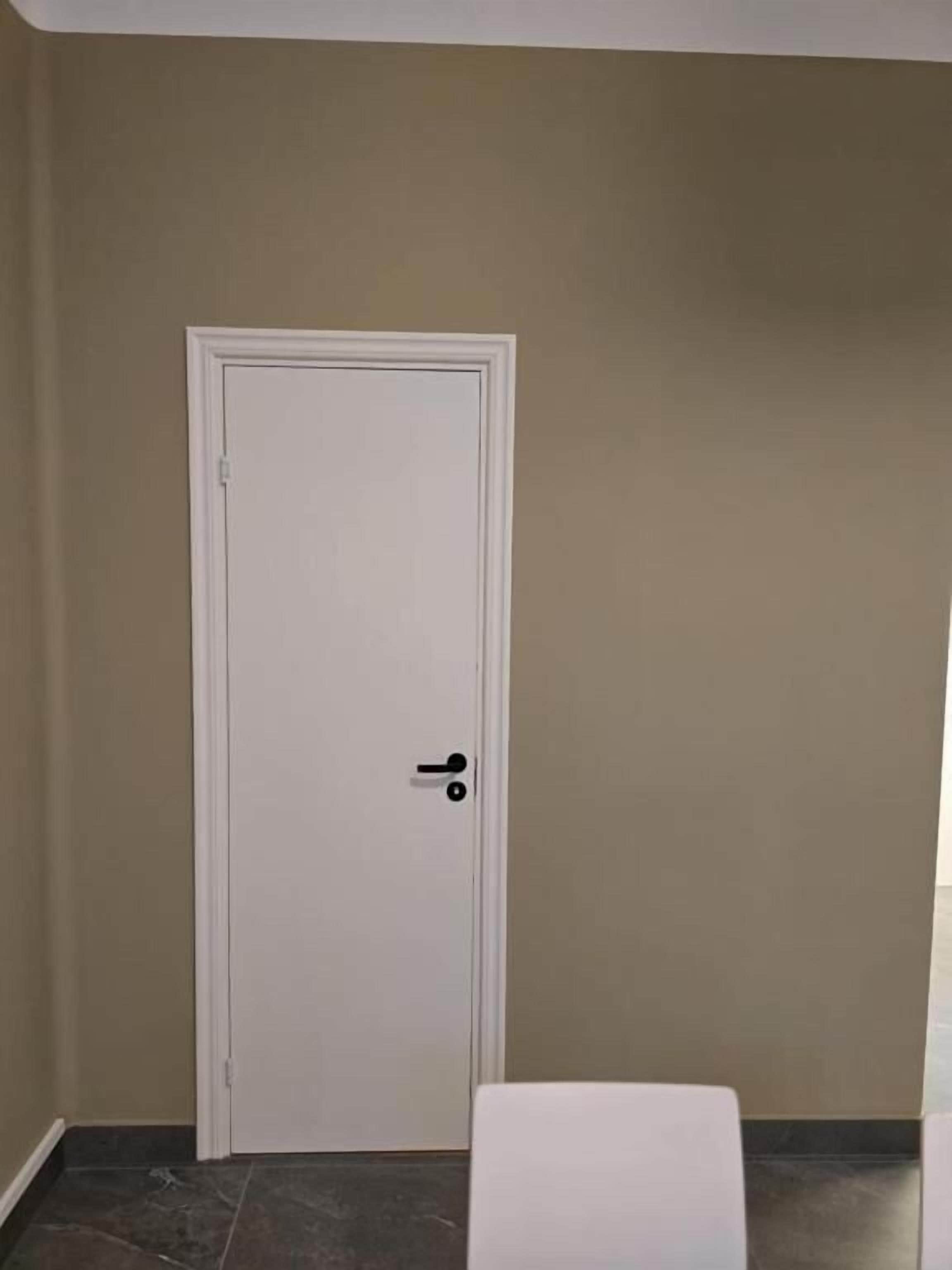 Une porte blanche fermée avec une poignée noire, un banc blanc et un mur de couleur beige.