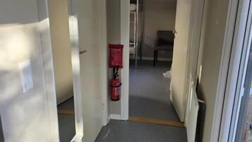 En smal hall med en brandsläckare på väggen, en matta vid ingången och en dörr som leder in till ett rum.