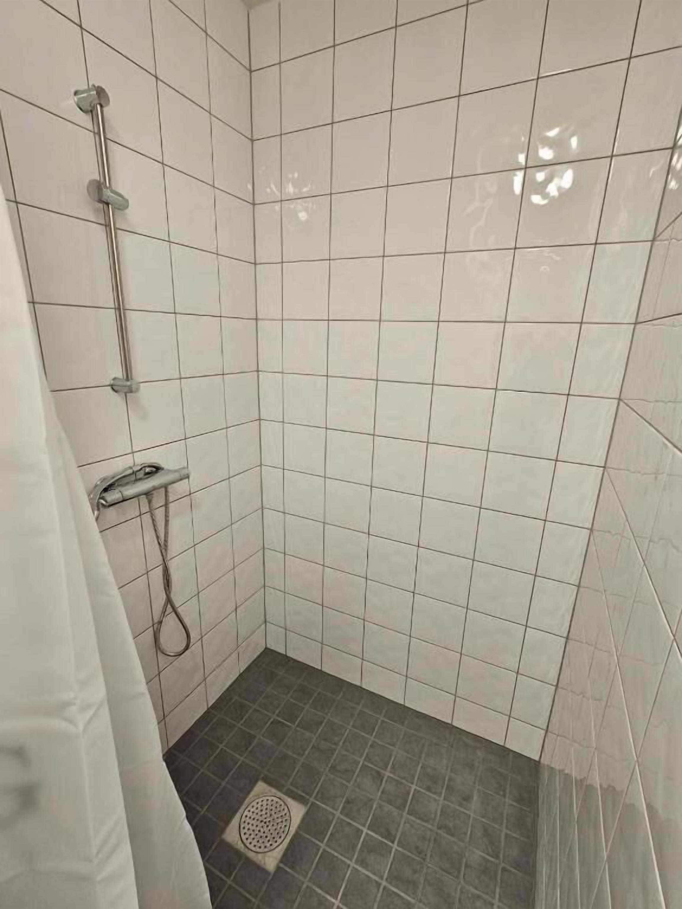Ett litet, kaklat duschutrymme med ett avlopp, ett duschmunstycke och ett handhållet duschmunstycke.