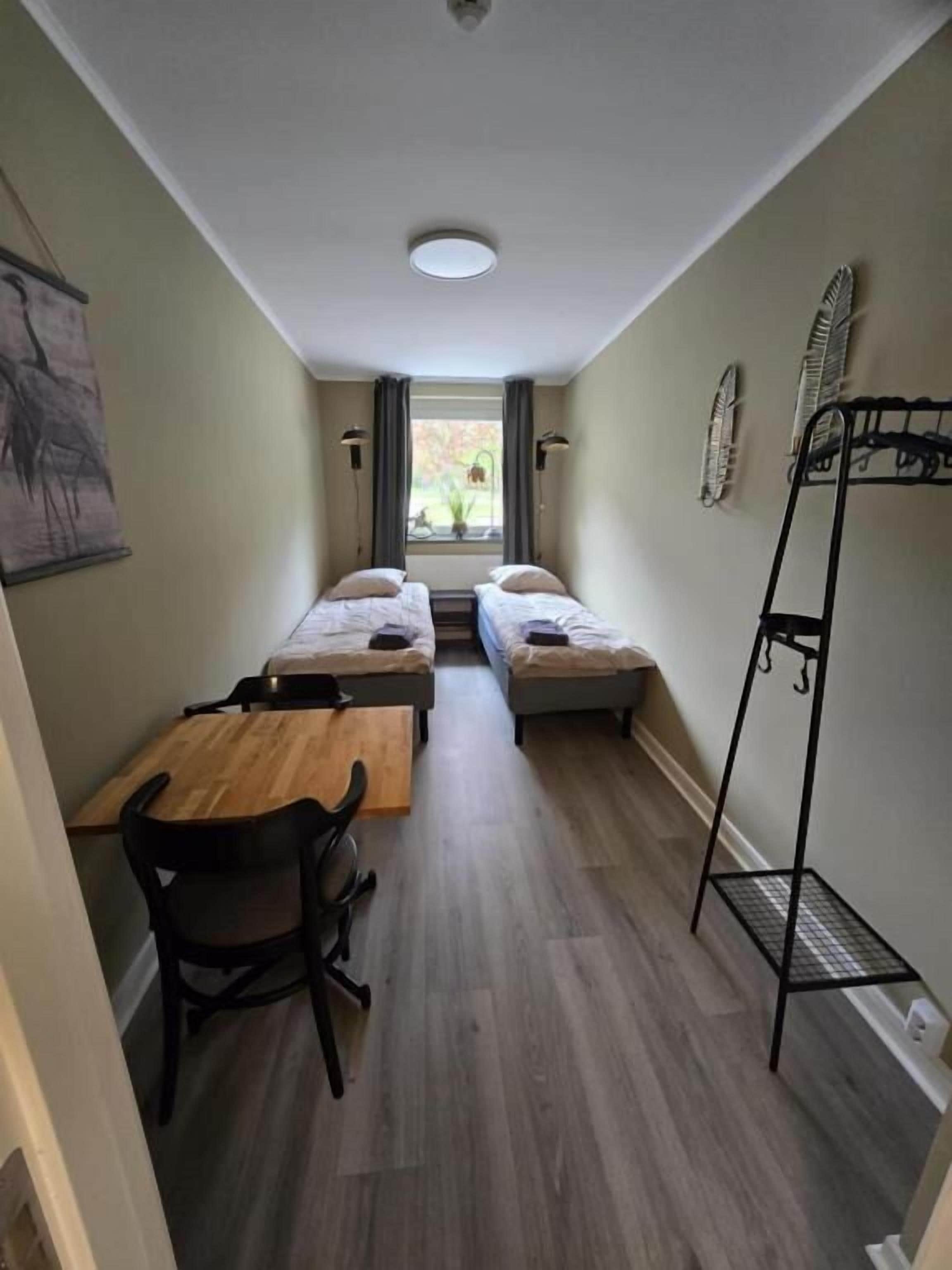 Une petite pièce avec deux lits simples, une table en bois, une chaise et un étagère en métal noir.