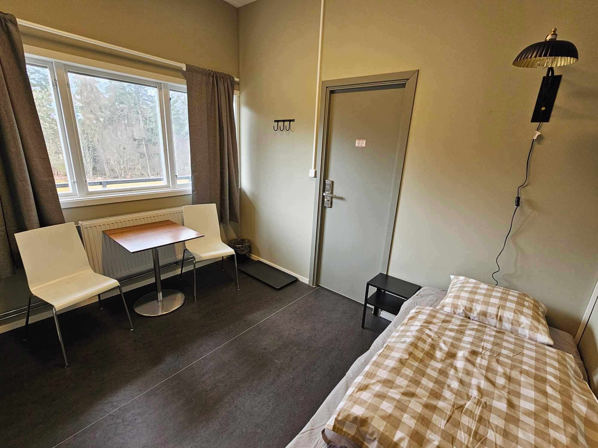 Um quarto pequeno e pouco mobiliado, com uma cama, uma mesa, duas cadeiras, um radiador, uma porta e uma janela.