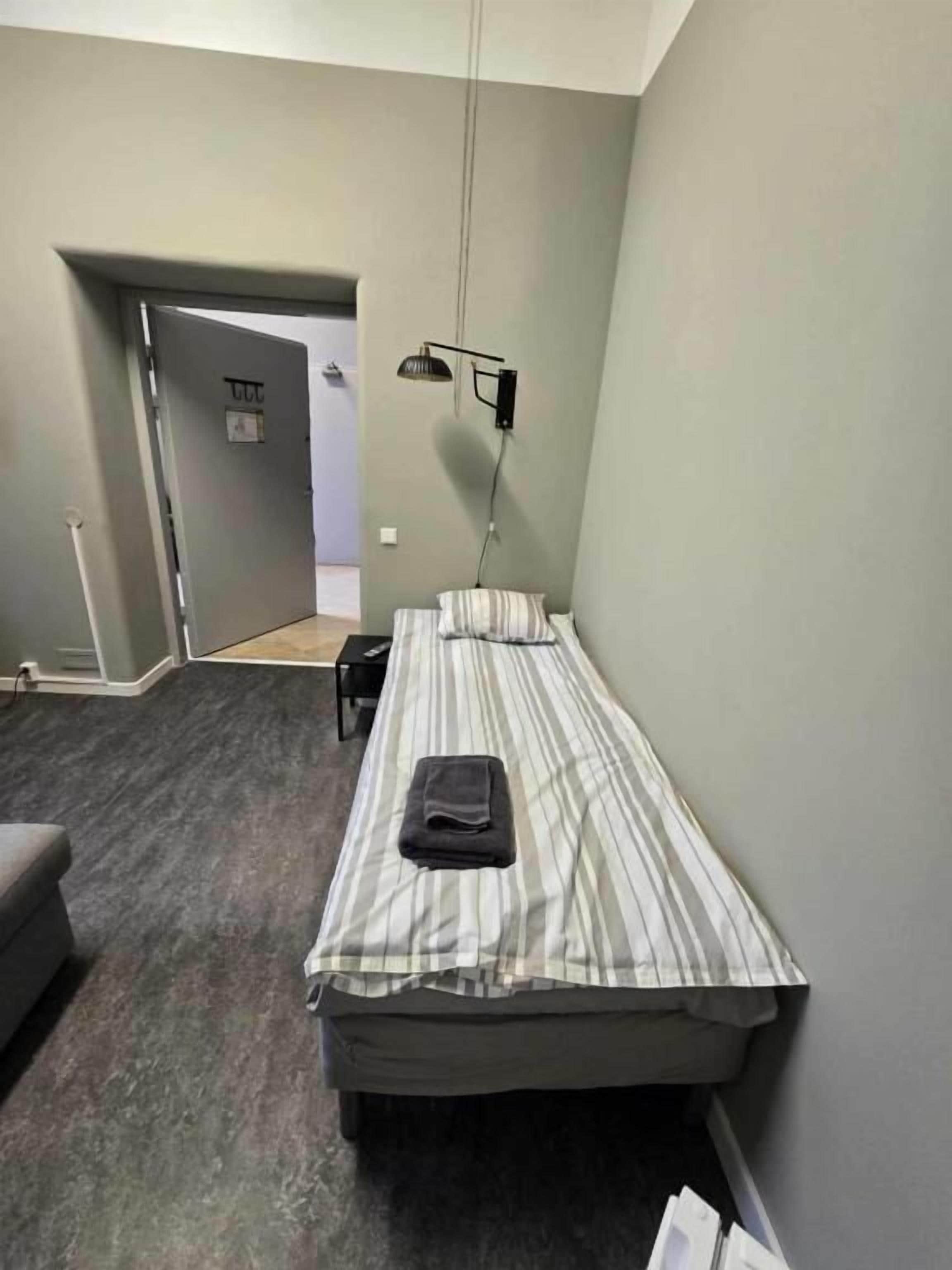 A single bed with a striped bedspread, a bedside lamp, and a dark-colored chair in a room with a grey carpet and walls.
