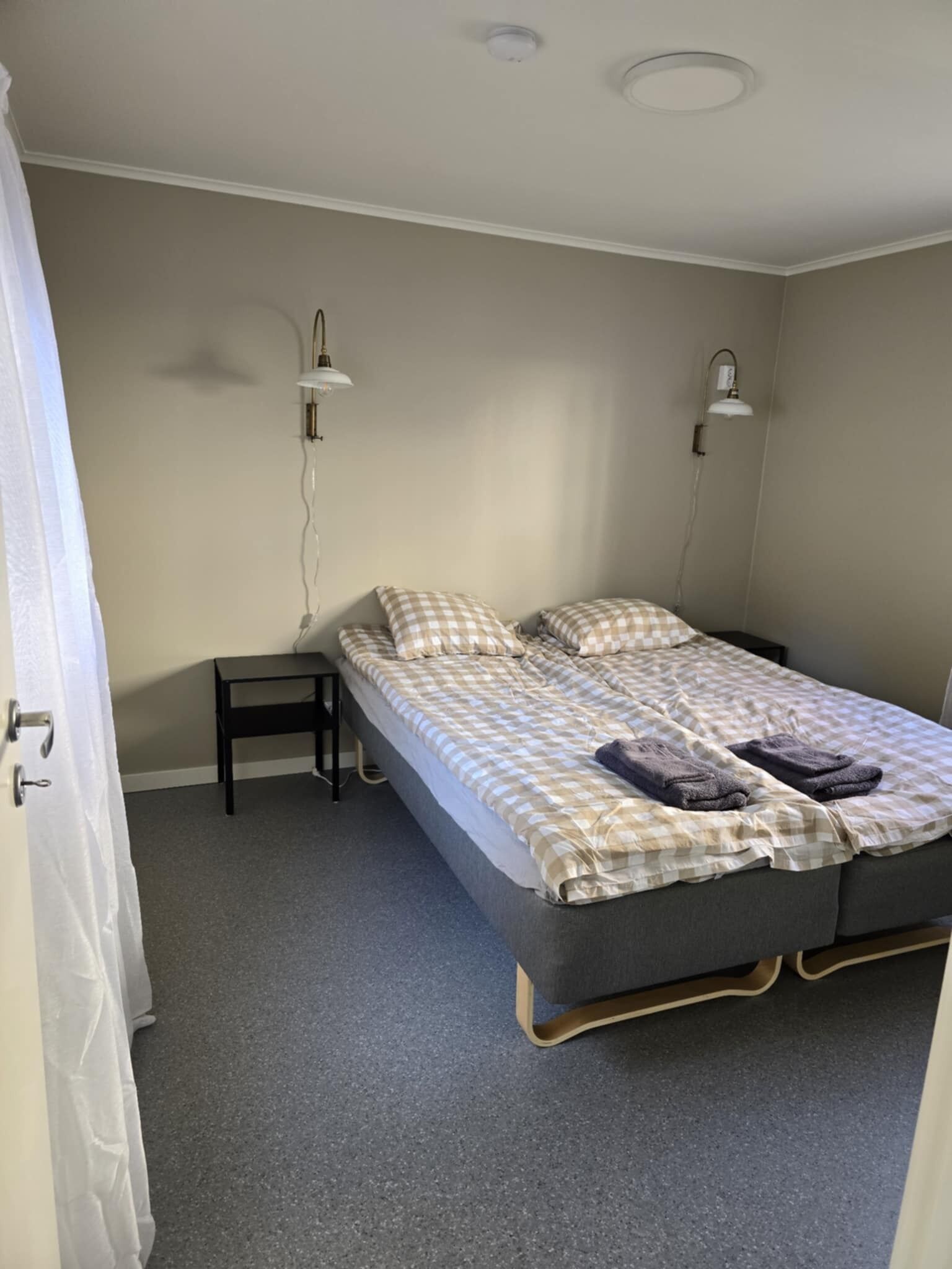 A single bed with a checkered duvet, two bedside lamps, a small table, and a grey carpeted floor.