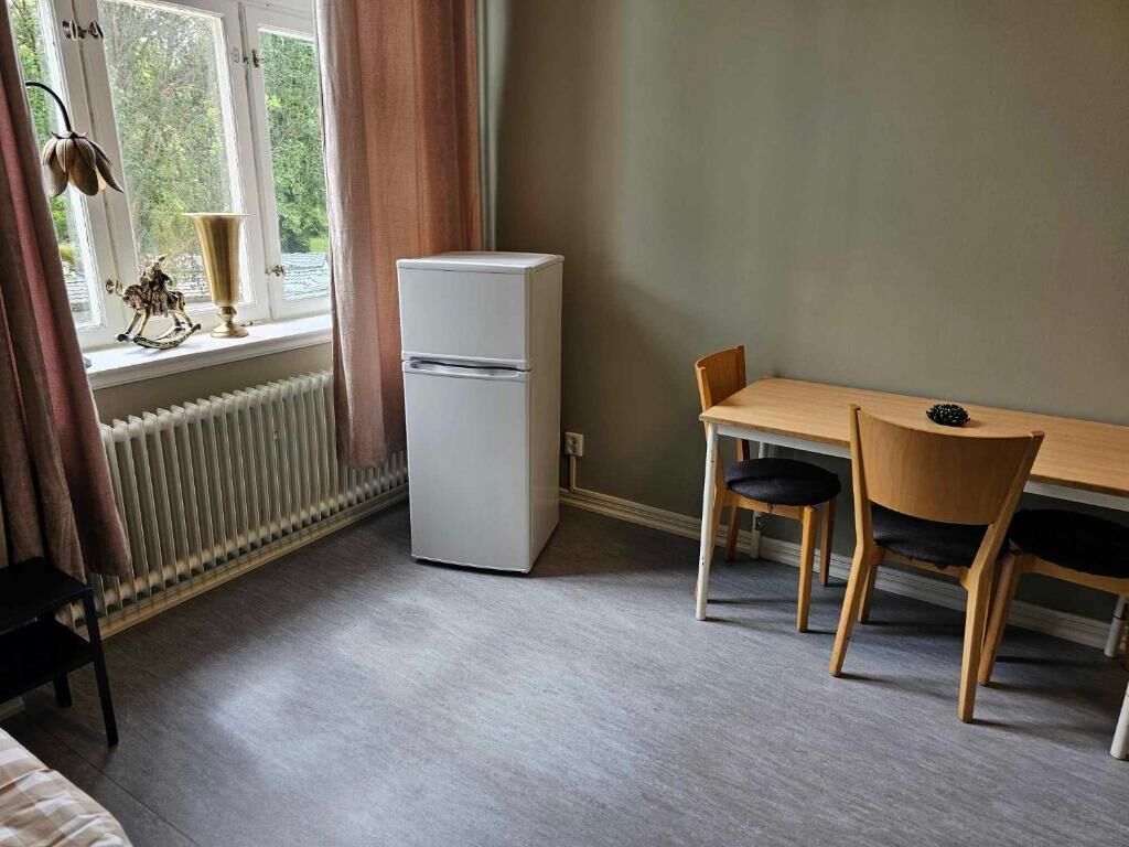 Ett rum med ett fönster, ett element, ett vitt kylskåp, ett träbord med två stolar och en säng.