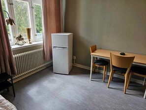 A room with a window, a radiator, a white refrigerator, a wooden table with two chairs, and a bed.