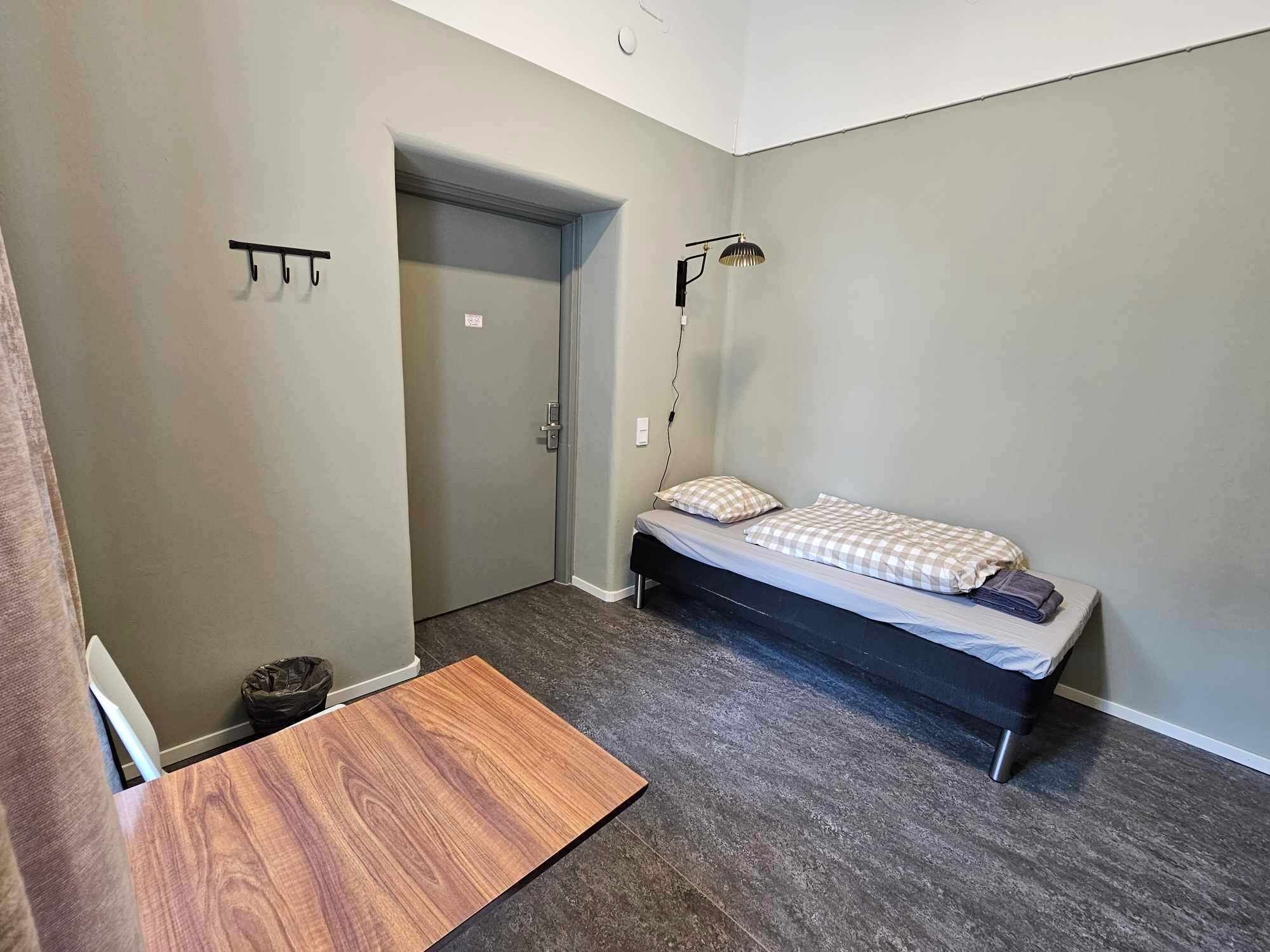 A small, modern bedroom with a single bed, a bedside table, a lamp, and a door leading to another room.