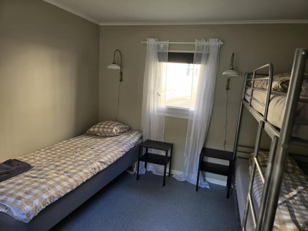 A single bed with a checkered bedspread, a small bedside table, a window with sheer curtains, and a bunk bed in the background.