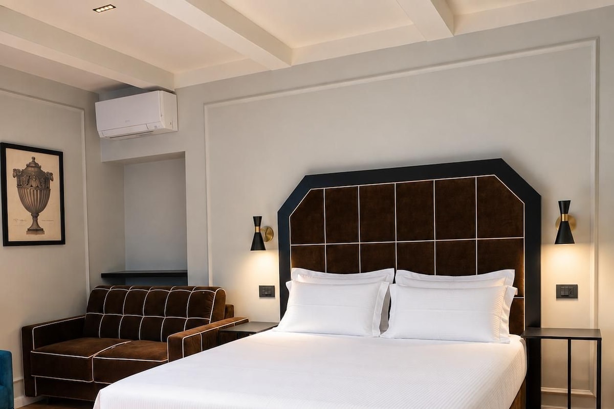 A hotel room with a large bed, a sofa, a chair, and a wall-mounted air conditioner.
