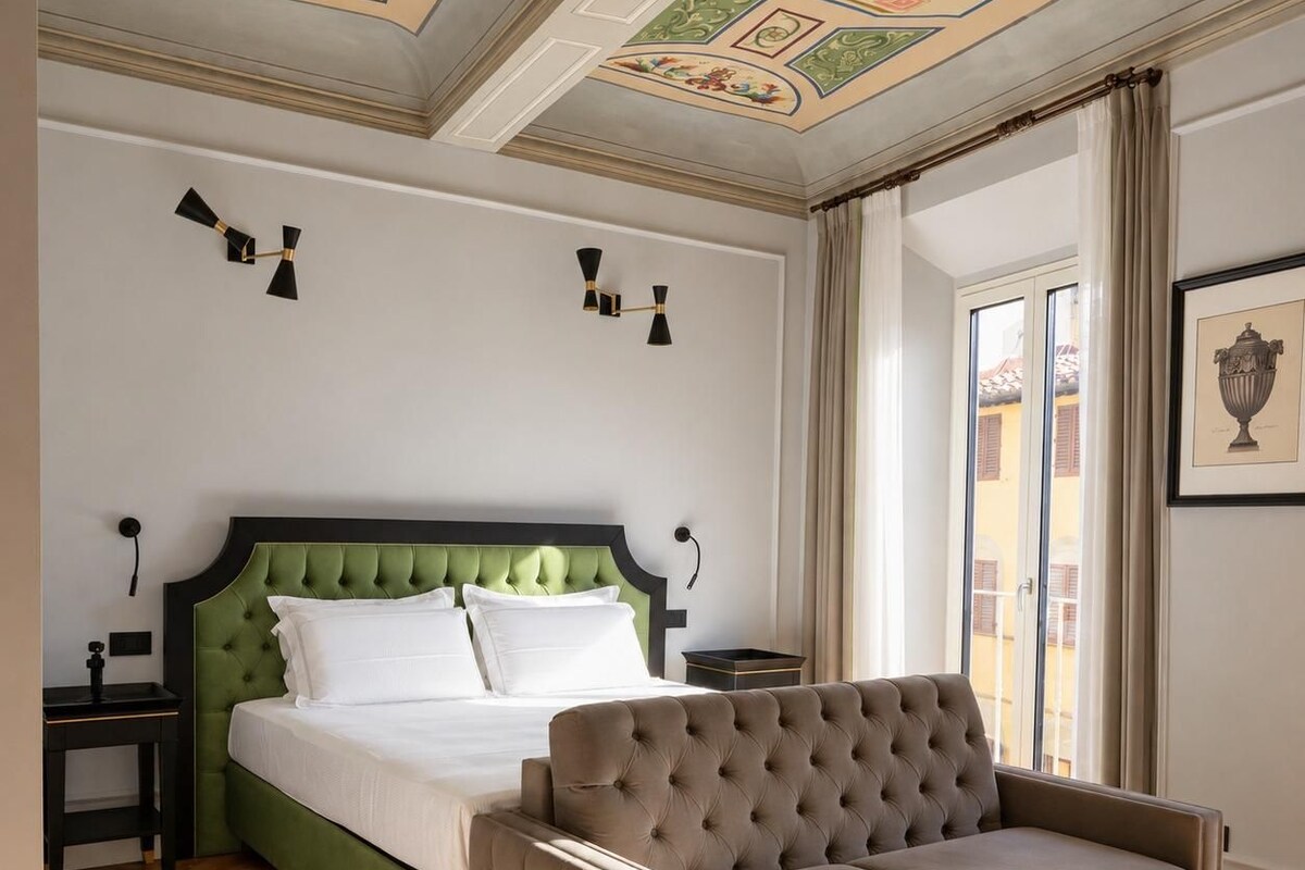 A bedroom with a green upholstered bed, a tufted armchair, a nightstand, and a wall-mounted lamp.