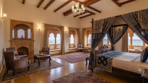 A spacious bedroom with a large bed, a sitting area with a chair and table, and a view of the sea through arched windows.