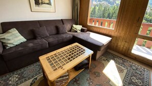 A cozy living room with a dark brown sofa, a wooden coffee table, and a large window offering a view of a forested area. - Studio for 4 people at the entrance to Méribel Mottaret (LES ALLUES)
