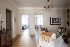 Ett rymligt vardagsrum med en vit soffa, en byrå i trä och en kristallkrona. Det finns två dörrar som leder till andra rum, och en inramad tavla på väggen.