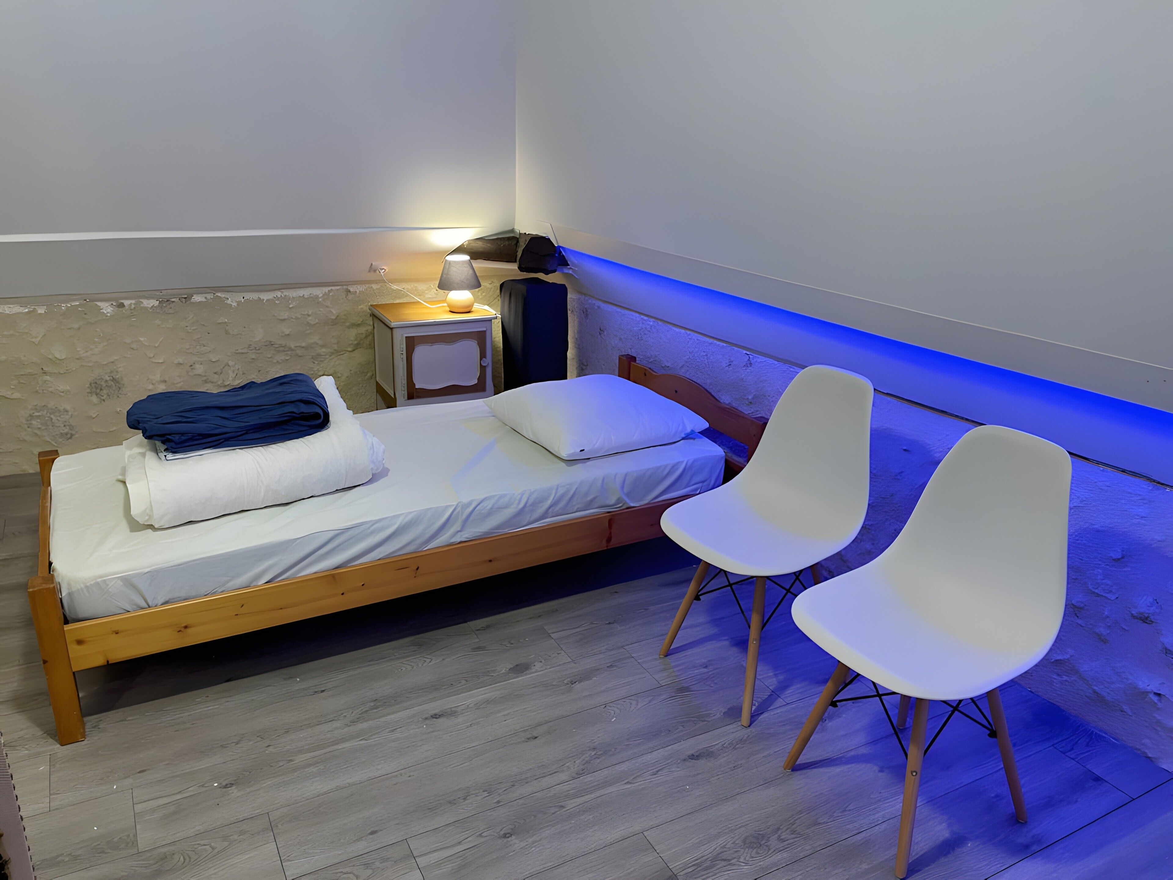 Uma cama de solteiro com uma mesa de cabeceira, duas cadeiras brancas e um abajur.