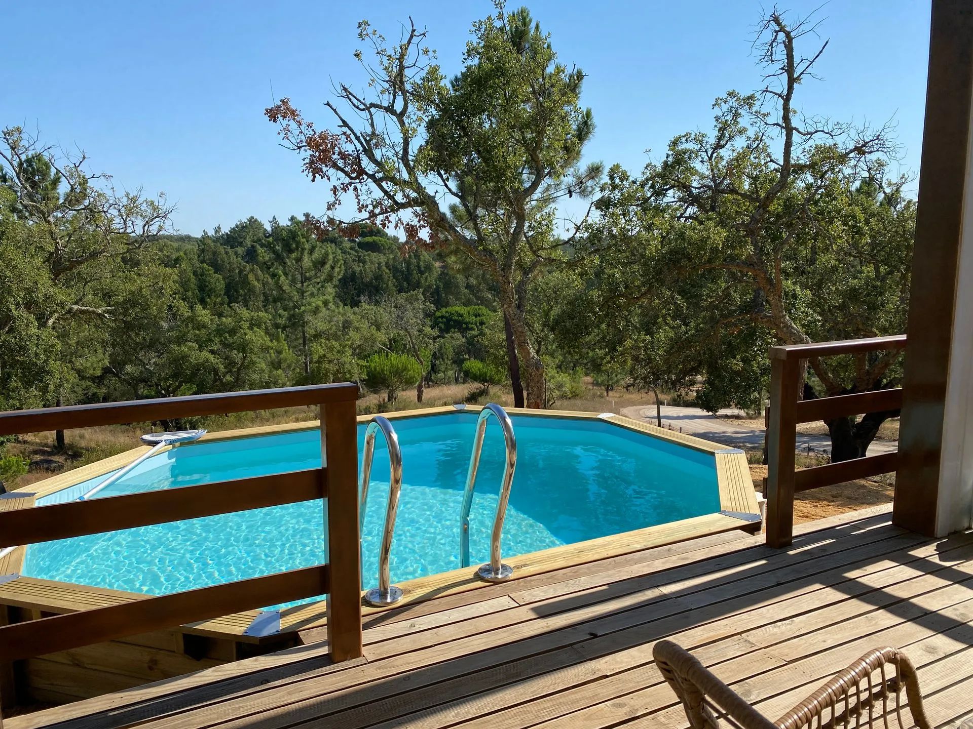 Una terraza de madera con piscina, rodeada de árboles y cielo despejado.