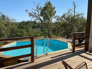 Una terraza de madera con piscina, rodeada de árboles y cielo despejado.