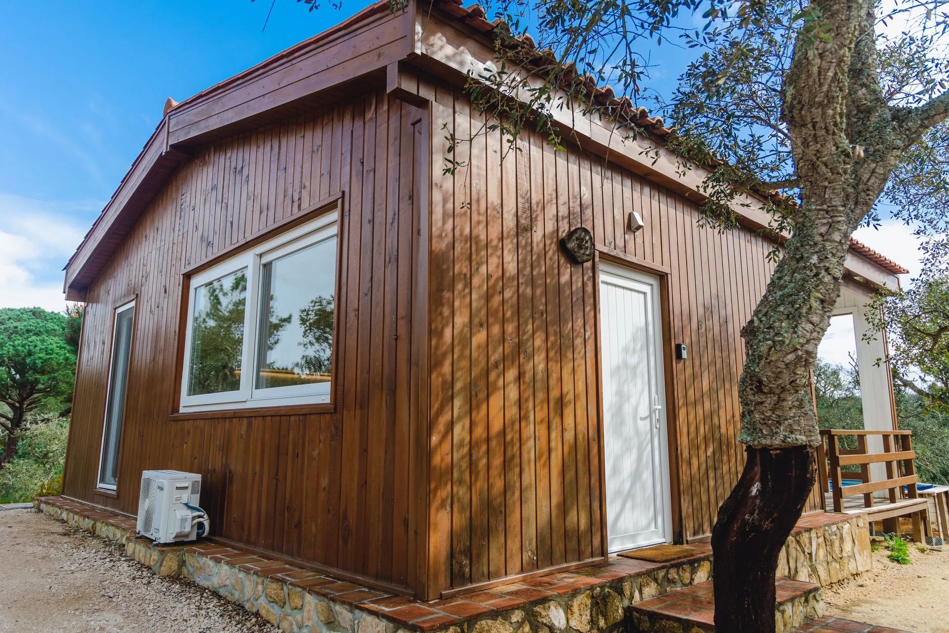 Una cabaña de madera con una sola puerta y ventanas, cimientos de piedra y un árbol en primer plano.