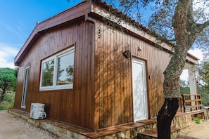 Una cabaña de madera con una sola puerta y ventanas, cimientos de piedra y un árbol en primer plano.
