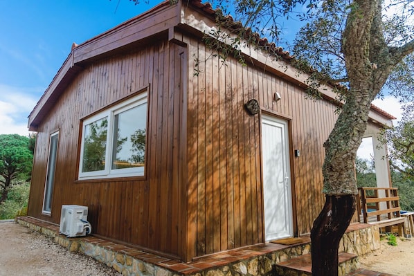 Una cabaña de madera con una sola puerta y ventanas, cimientos de piedra y un árbol en primer plano.