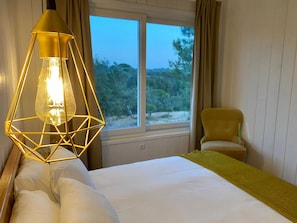 Une chambre d’hôtel avec un lit, une chaise, une fenêtre donnant sur la verdure et un luminaire au design géométrique original.