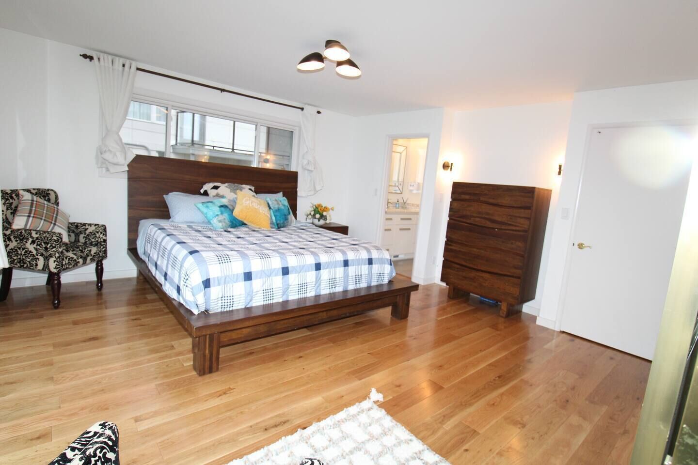 A bedroom with a wooden bed, a chair, a dresser, and a window with curtains.