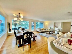 Uma sala de estar espaçosa com mesa de jantar, cadeiras e sofá. Há uma janela grande que proporciona luz natural e vista para o exterior.