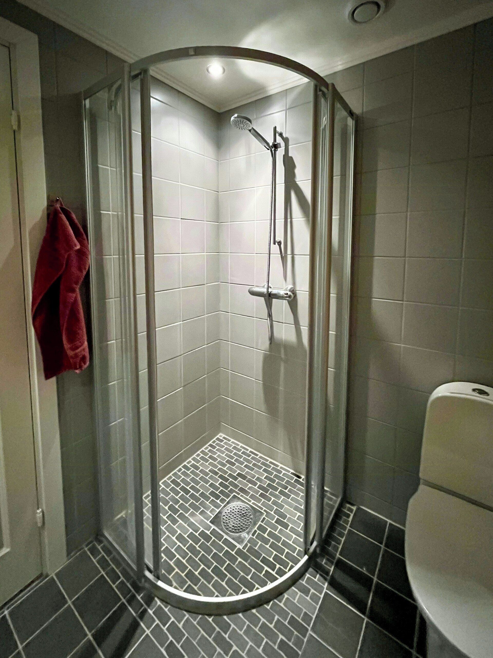 Een douche in hoekopstelling met een glazen deur, een betegelde vloer, en een rode handdoek aan de muur.