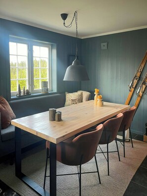 A dining area with a wooden table, four chairs, a hanging lamp, and a window with a view of trees.