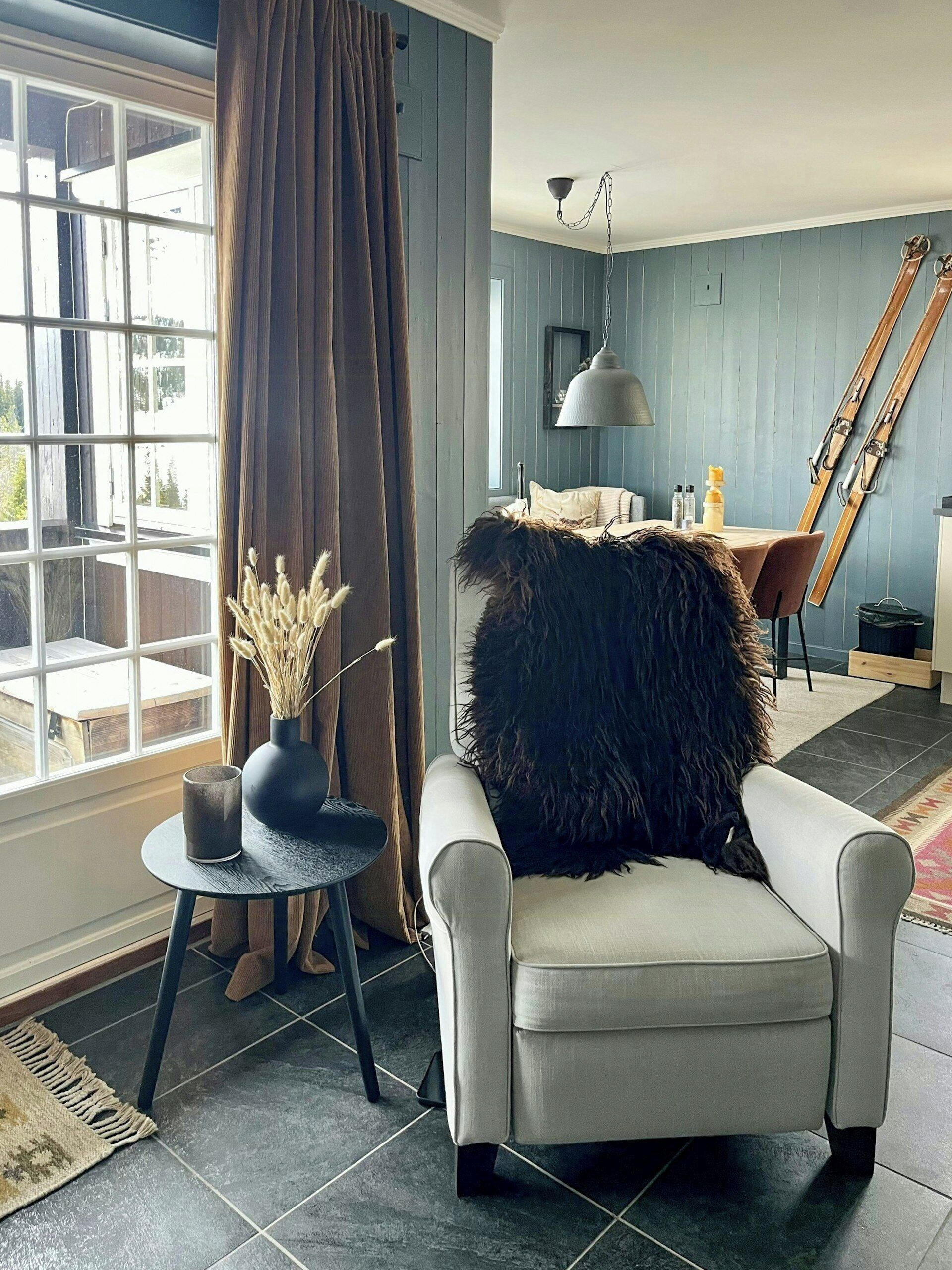 A room with a large window, a chair with a fur throw, a small table with a vase, and a dining area with a table and chairs.