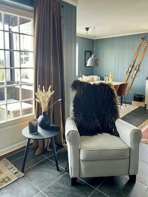 Ein Zimmer mit einem großen Fenster, einem Sessel mit einem Fellüberwurf, einem kleinen Tisch mit einer Vase und einem Essbereich mit Tisch und Stühlen.