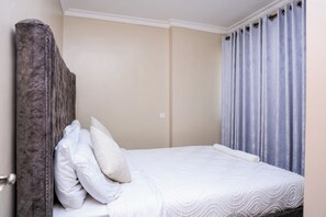 A single bed with white bedding and pillows, a wooden headboard, and a window with curtains. - 1 Bedroom Apartment in Utawala Nairobi  Near JKIA Free Wi-Fi & Parking (Nairobi)