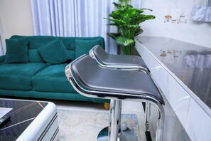 A modern kitchen with a white countertop, chrome bar stools, and a green sofa in the background. - 1 Bedroom Apartment in Utawala Nairobi  Near JKIA Free Wi-Fi & Parking (Nairobi)