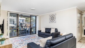A modern living room with a black leather sofa, a coffee table, and a patterned rug. There is a sliding glass door leading to an outdoor area with a view of buildings and trees. - The Poplars Apartment with views and  Swimming Pool (Nelson Bay)