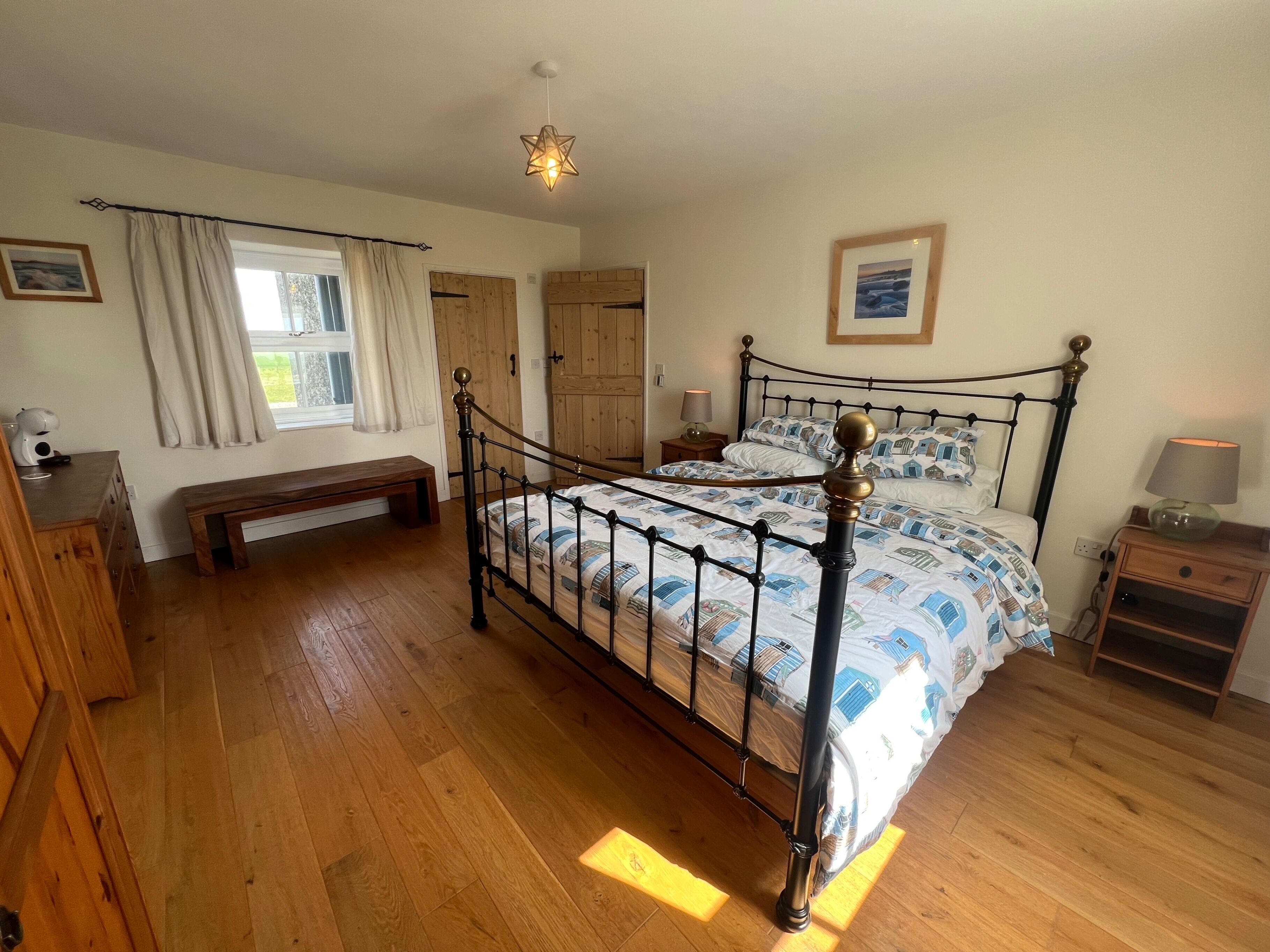 A bedroom with a metal-framed bed, wooden flooring, a bench, a bedside table with a lamp, and a window with curtains.