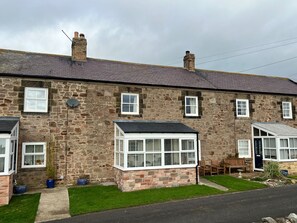 A stone house with multiple windows, a chimney, and a small garden area with a bench and potted plants. - Cottage - Sleeps 5 - Parking & Wood Burning Stove (Alnwick)