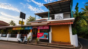 Un edificio de dos pisos con un letrero que indica Hotel Candolim ALOFT, un restaurante con diversas opciones de cocina india y una moto estacionada.