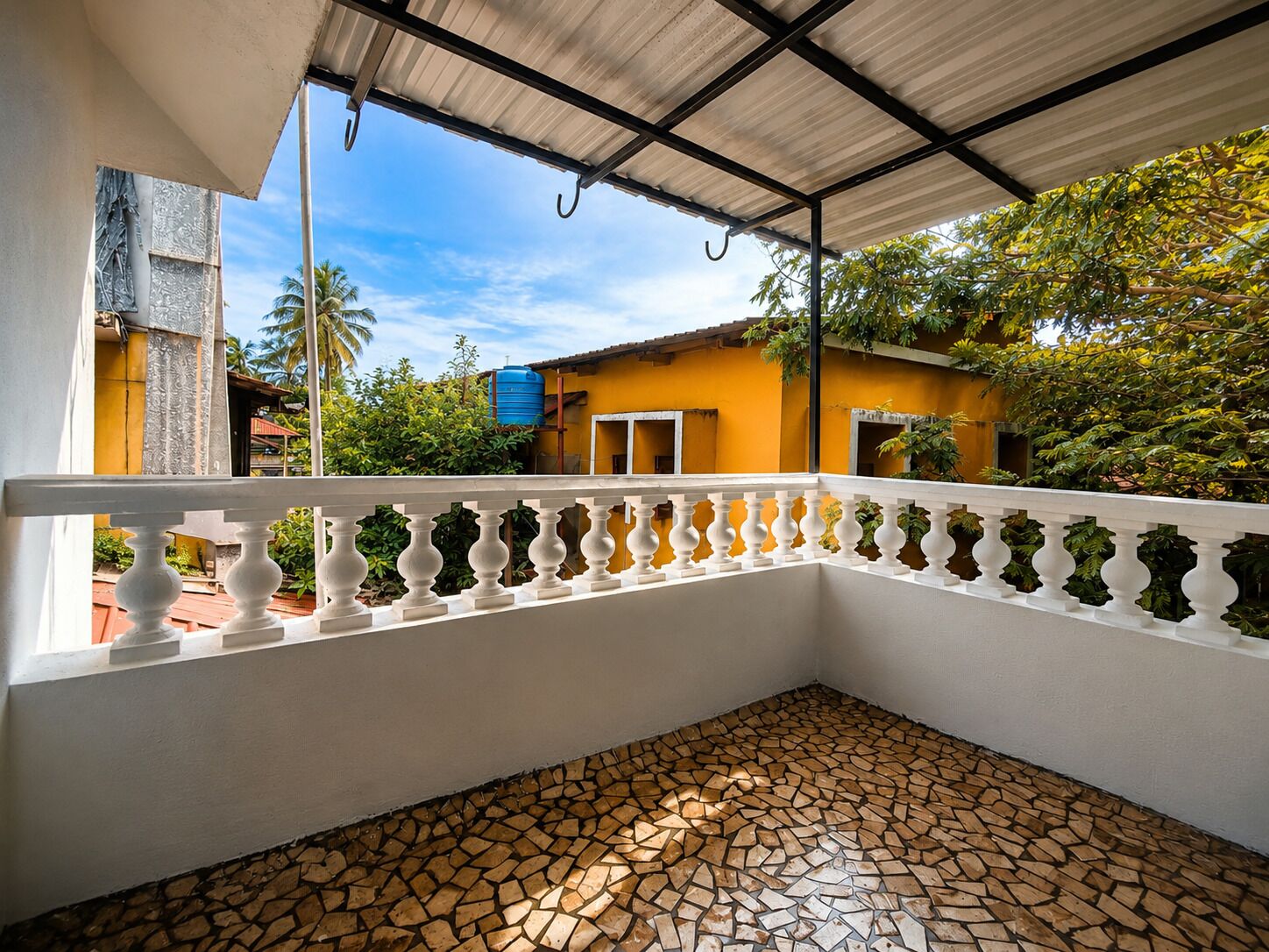 Un balcon couvert avec une balustrade blanche, un sol carrelé et une vue sur un immeuble jaune et de la verdure.