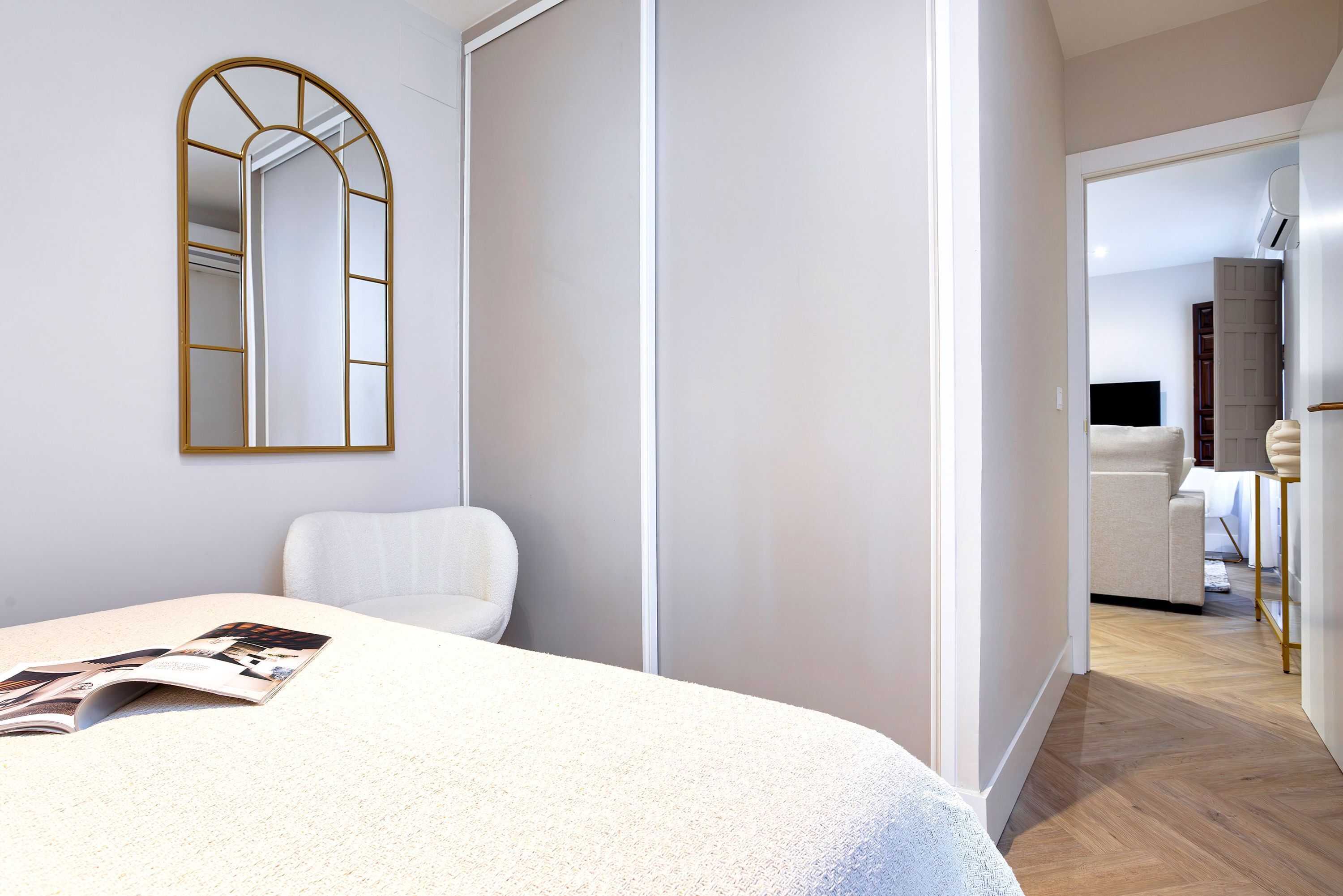 A bedroom with a bed, a white chair, a mirror, and a door leading to a bathroom.