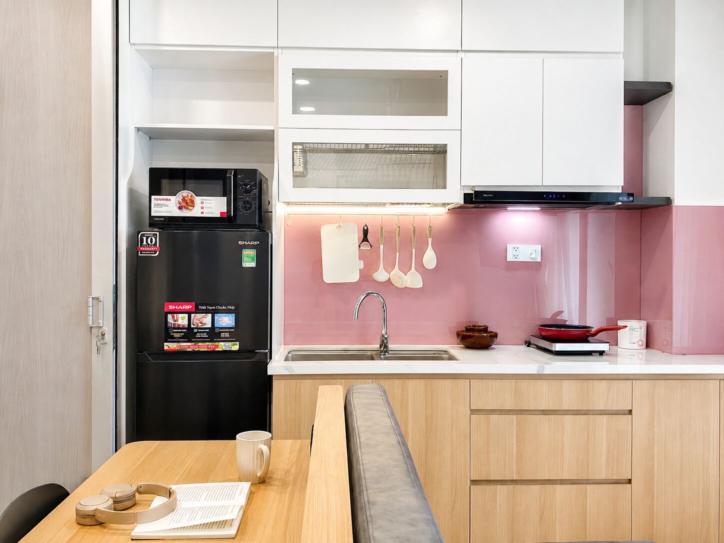 A compact kitchen with a refrigerator, microwave, and stove. There is a sink with hanging utensils and a table with a mug and headphones.