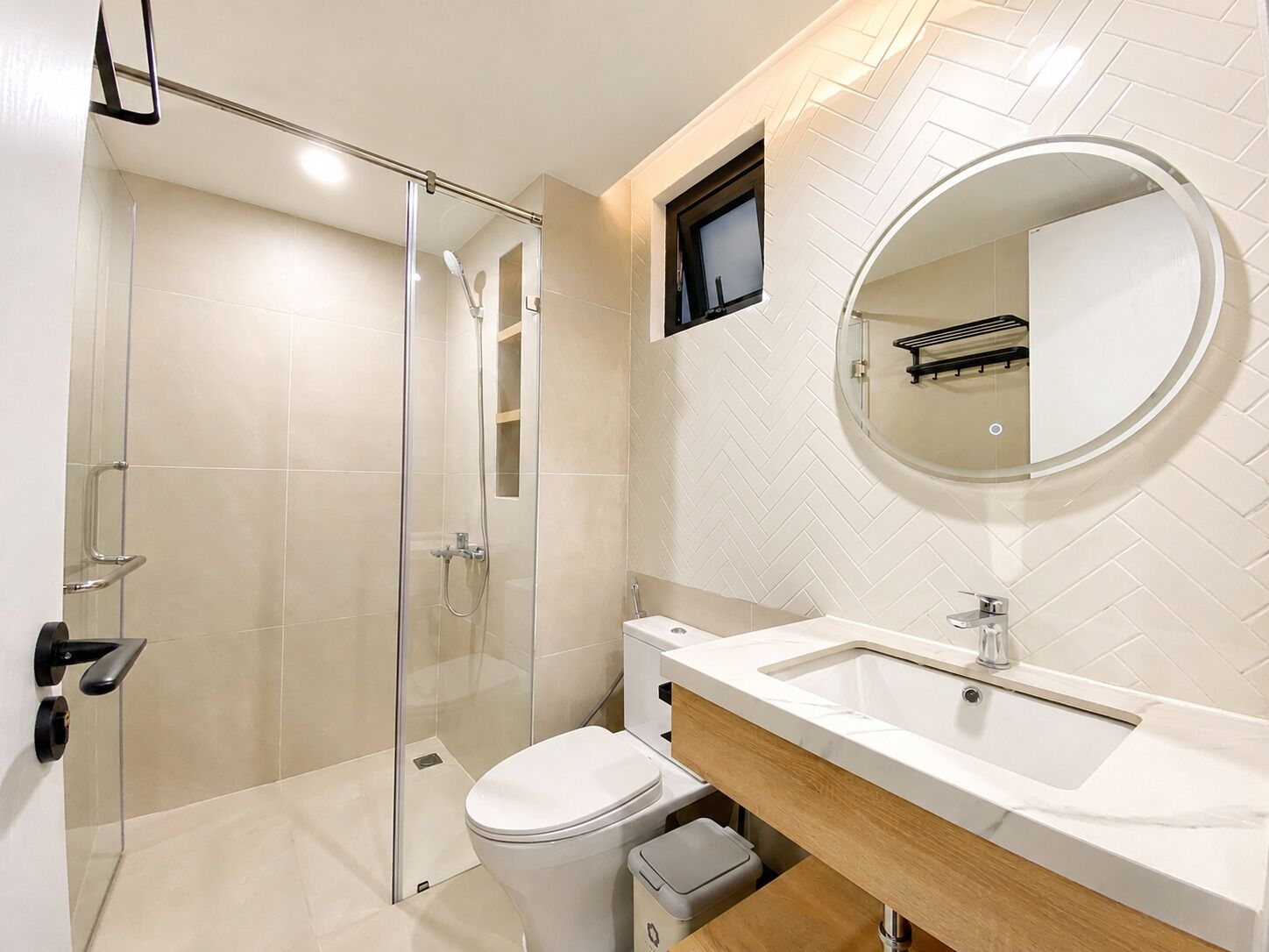 A modern bathroom with a glass shower enclosure, a toilet, a sink with a mirror, and a small window.