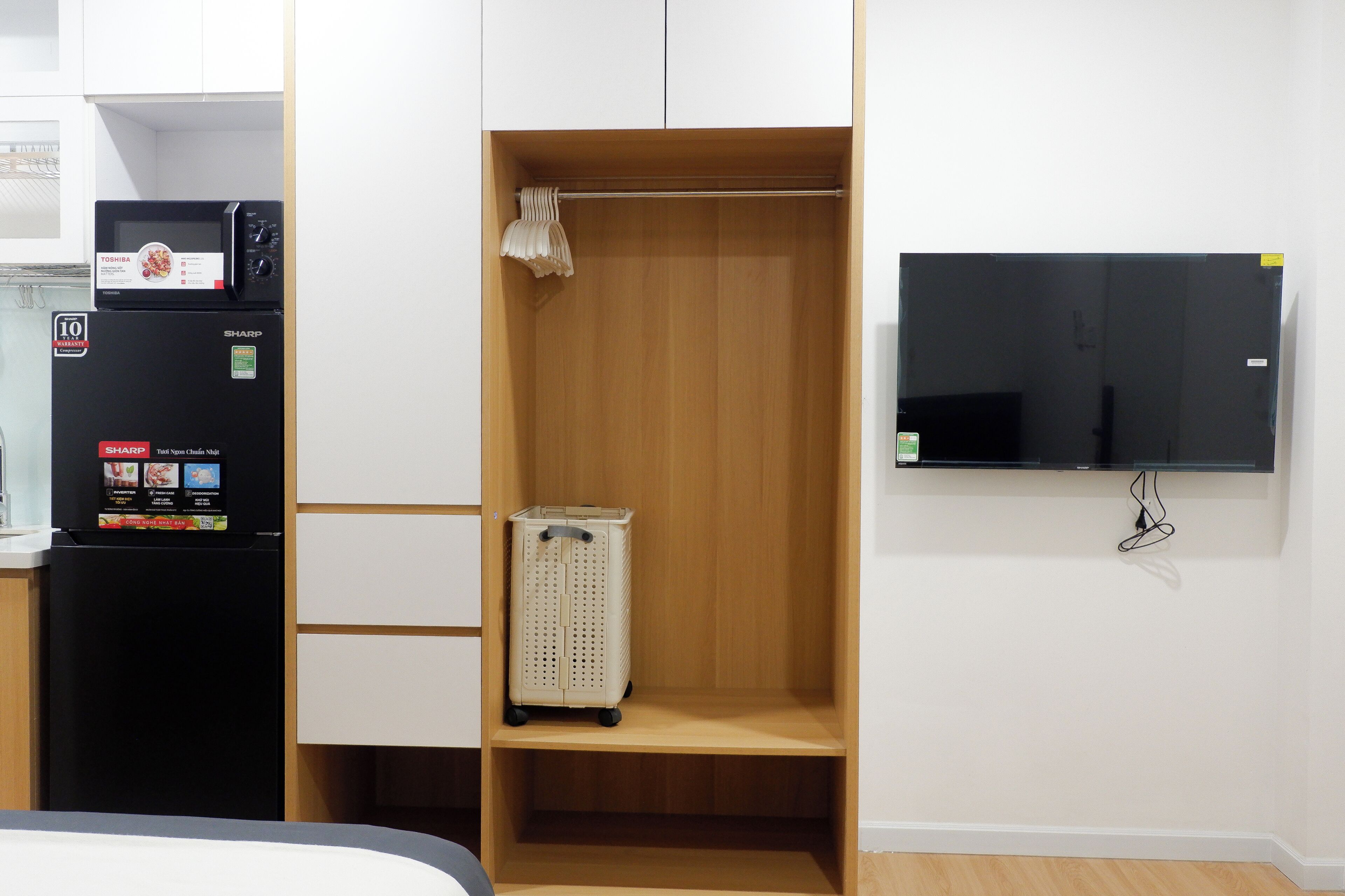 A compact room with a black refrigerator, a television mounted on the wall, and a wooden wardrobe with a hanging rack.