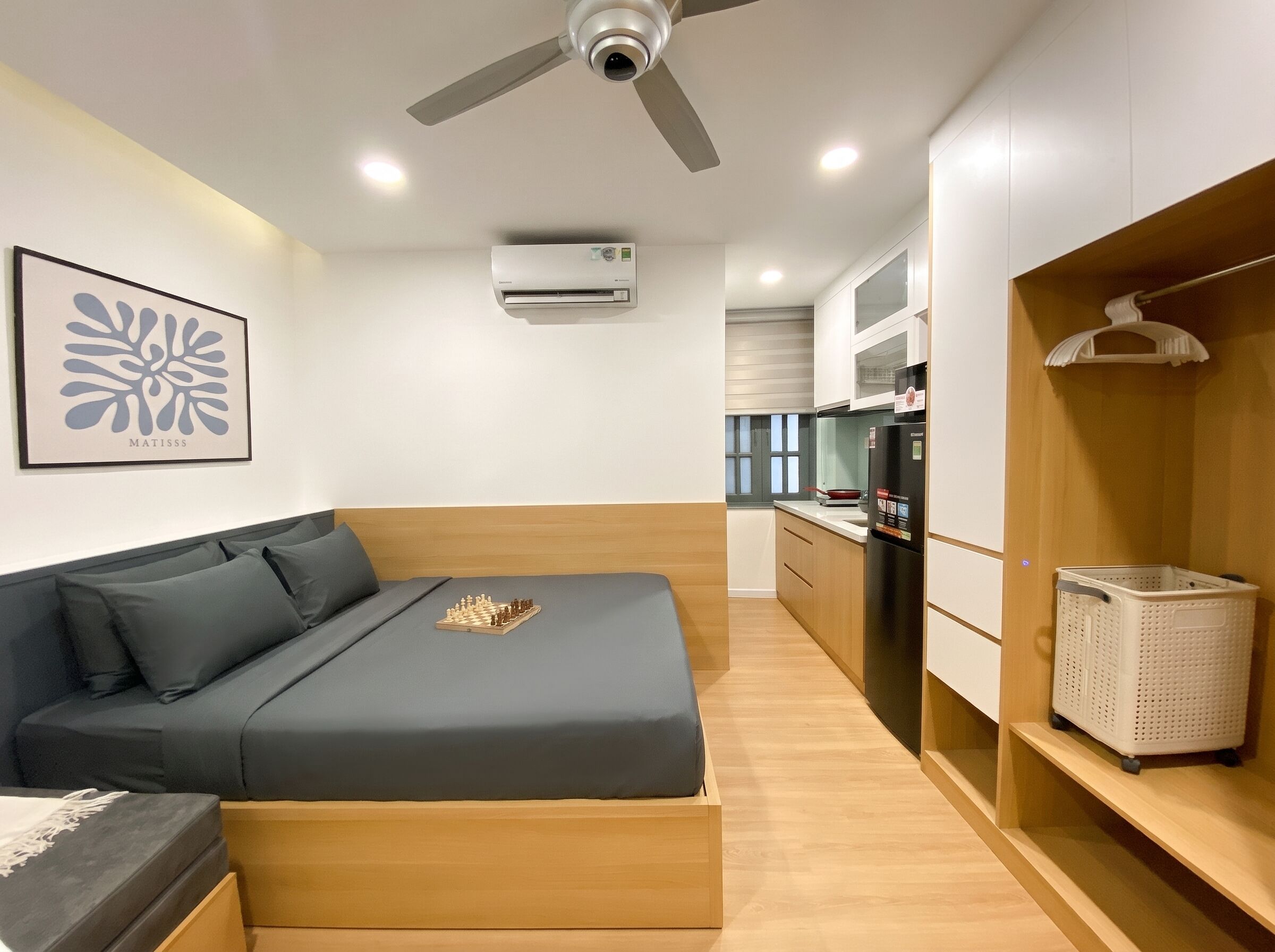 Un dormitorio moderno con cama, ventilador de techo, aire acondicionado y una zona de cocina con refrigerador y pileta.