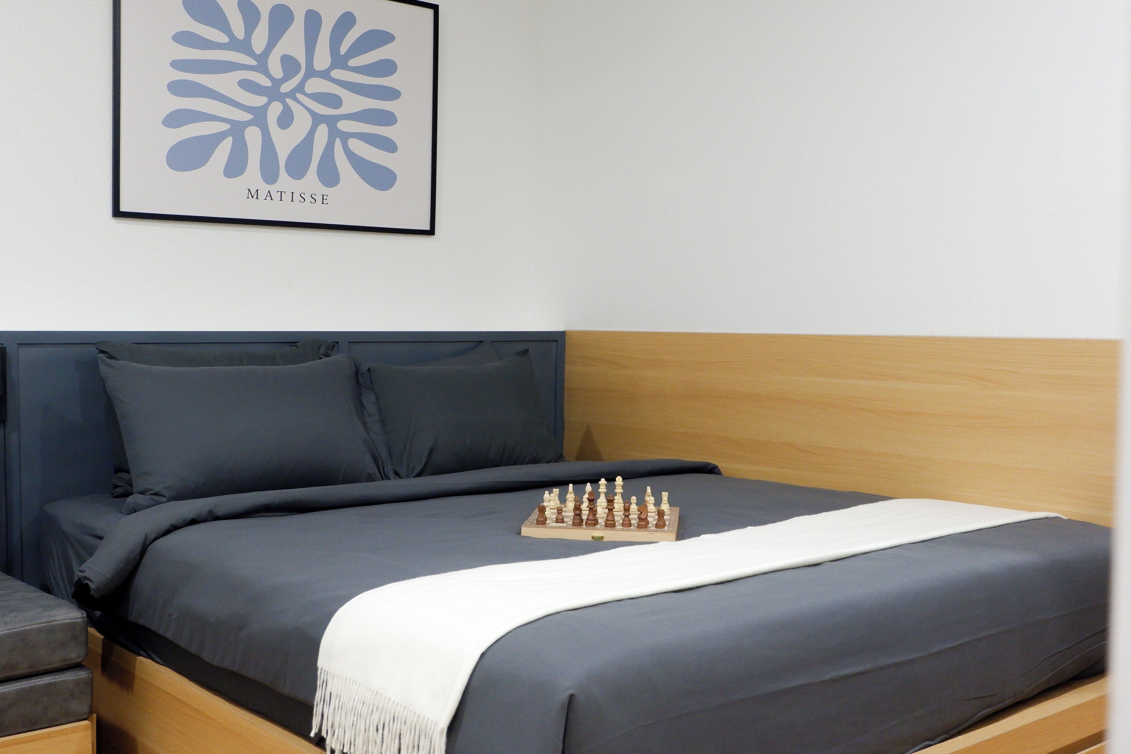 A neatly made bed with a chessboard on it, a framed artwork on the wall, and a bedside table with a lamp.