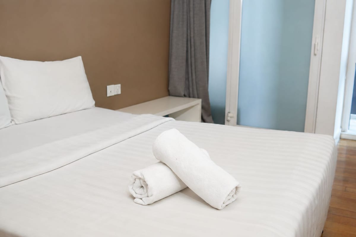 A neatly made bed with a rolled white towel, a bedside table, and a window with curtains.